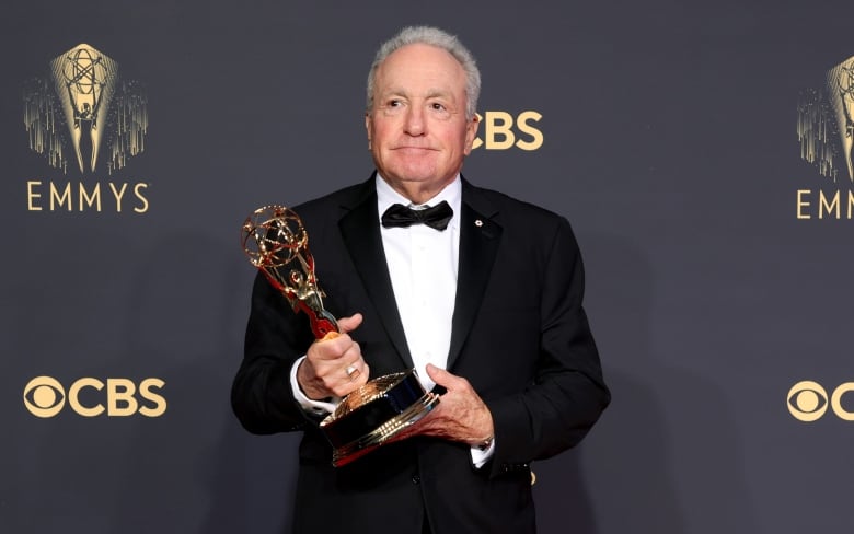 A white-haired man wearing a black tuxedo holds an Emmy Award statue.