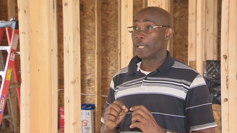 A Black man wearing glasses  and a black and white stripped shirt standing in an unfinished house