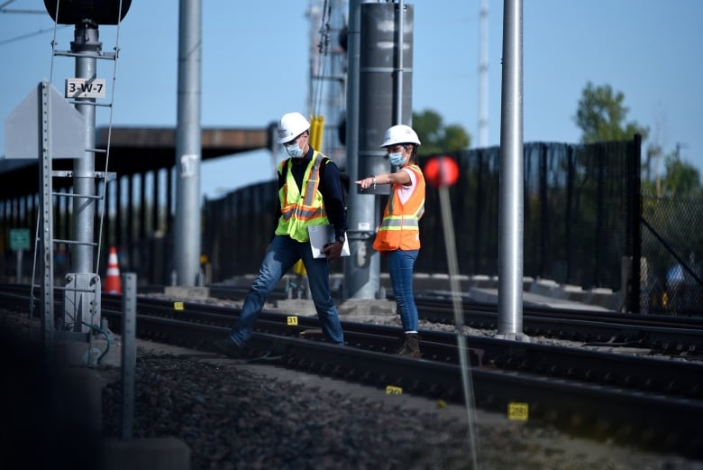 A person in a hardhat and orange vest points with her arm and a man in a hard hat and a yellow vest walks in that direction over train tracks.