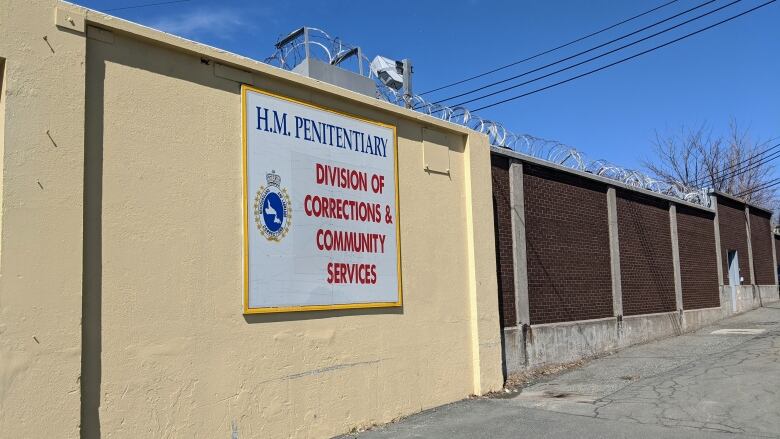 The front entrance of Her Majesty's Penitentiary in St. John's, NL. The prison sign is hung on a pale yellow wall, bordering a brick wall with barbed wire on top.