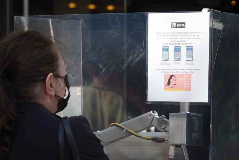 A person in a protective facemask reads a notification about vaccine card and mask requirements outside of a restaurant, mounted on a glass wall.