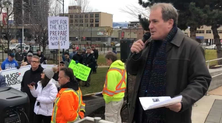 A photo of a middle aged man holding a microphone and speaking to a rally of several dozen people at a public square in Kelowna, B.C. 