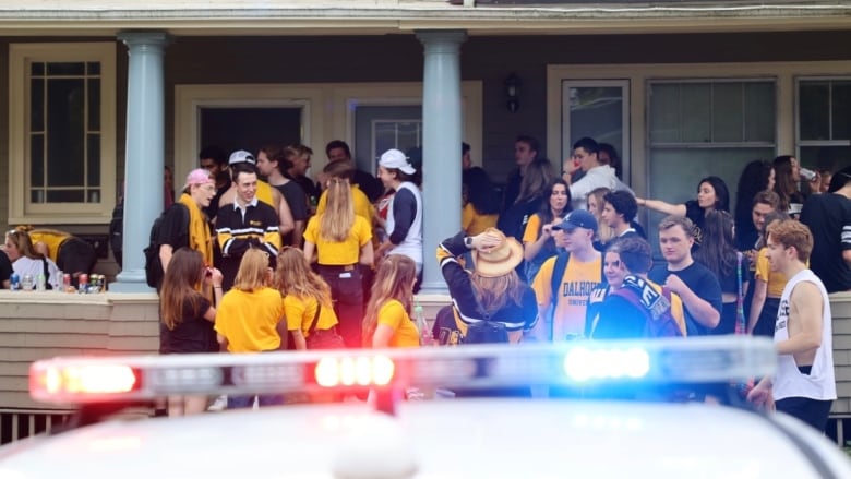 A crowd of young adults stand in front of a house. The rooftop lights of a police cruiser flash in the foreground.