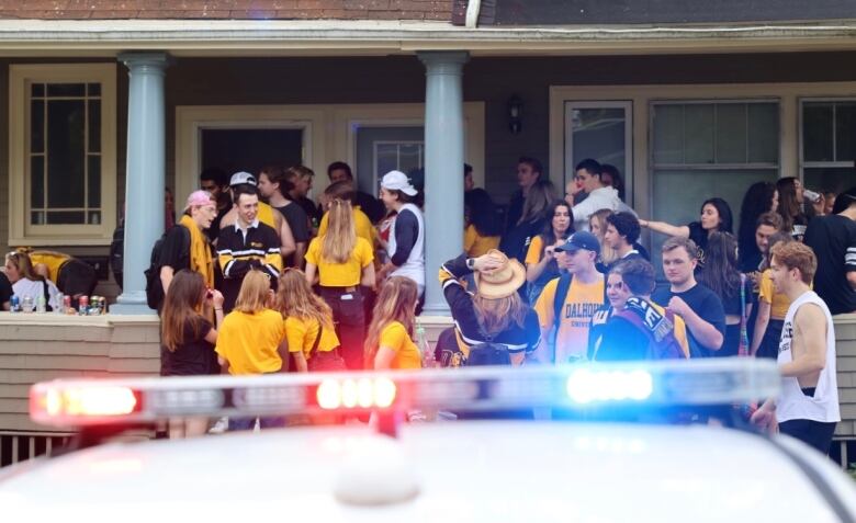 A crowd of young adults stand in front of a house. The rooftop lights of a police cruiser flash in the foreground.