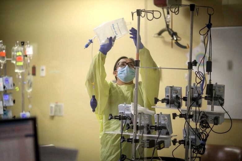 A health-care worker wearing a blue medical mask, yellow gown and blue surgical gloves, replaces an IV bag of fluid on a metal pole overhead.