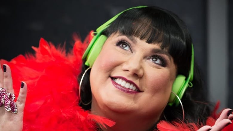 A woman with black bangs and long hair wearing a red boa on their shoulders and neon green sunglasses on top of her head. She sits in front of a mic in a radio studio. 