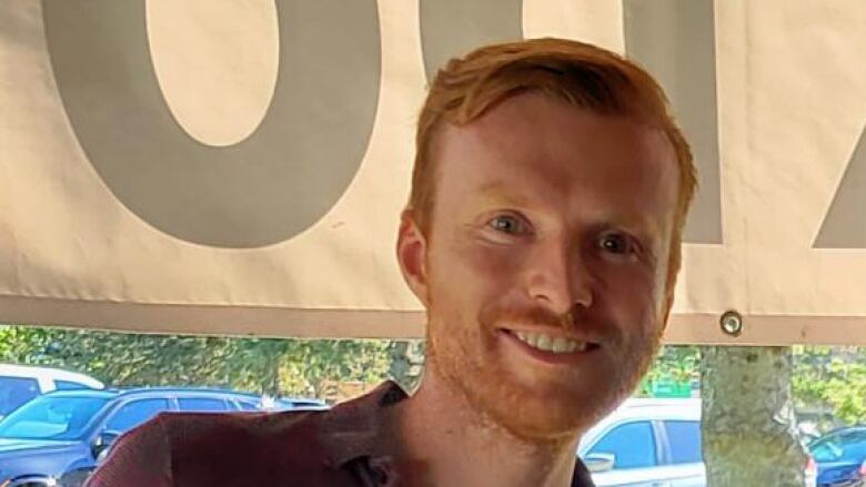 A man with red hair and beard smiles while standing outside.