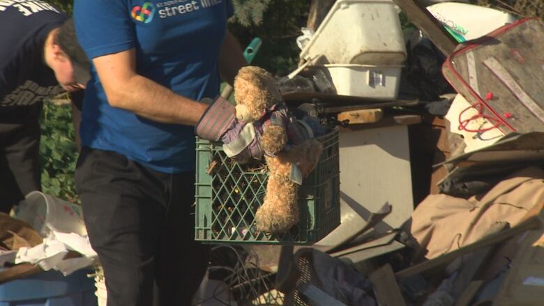 A person is carrying a crate full of items, including a teddy bear. Debris can be seen in the background.