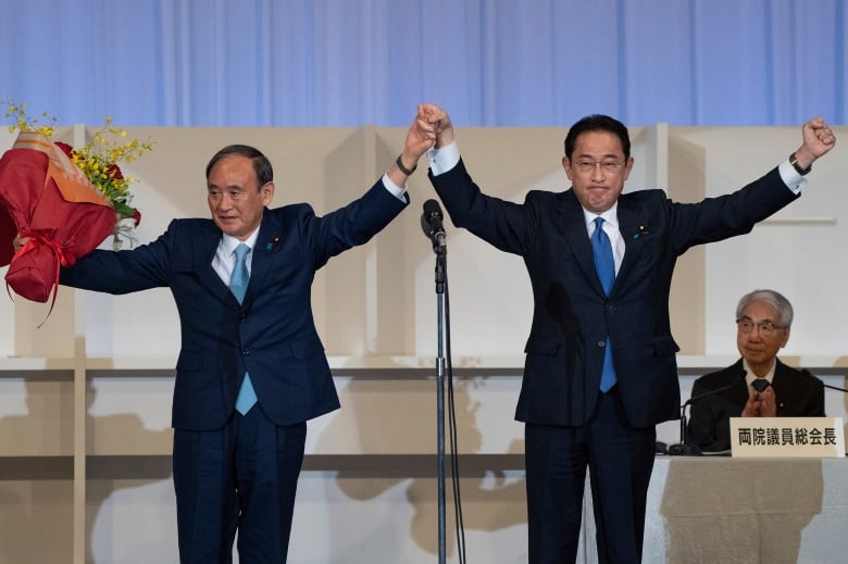 Two men stand on a stage, one holding a bouquet of flowers, holding hands and raising their arms.