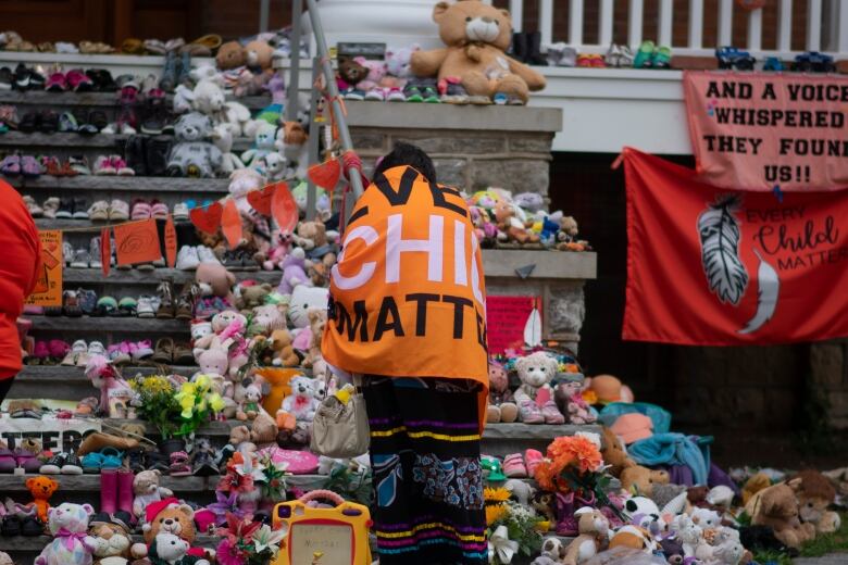 A person stands in front of steps filled with children's toys and clothes. The person is wrapped in a banner.