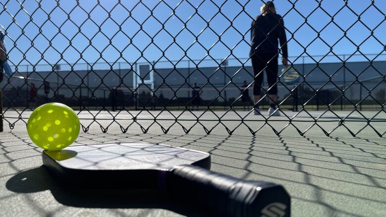 A pickleball paddle and ball are seen in the foreground in front of a chain link fence with a court behind where a game is underway.