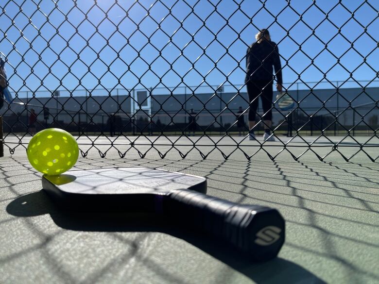 A pickleball paddle and ball are seen in the foreground in front of a chain link fence with a court behind where a game is underway.