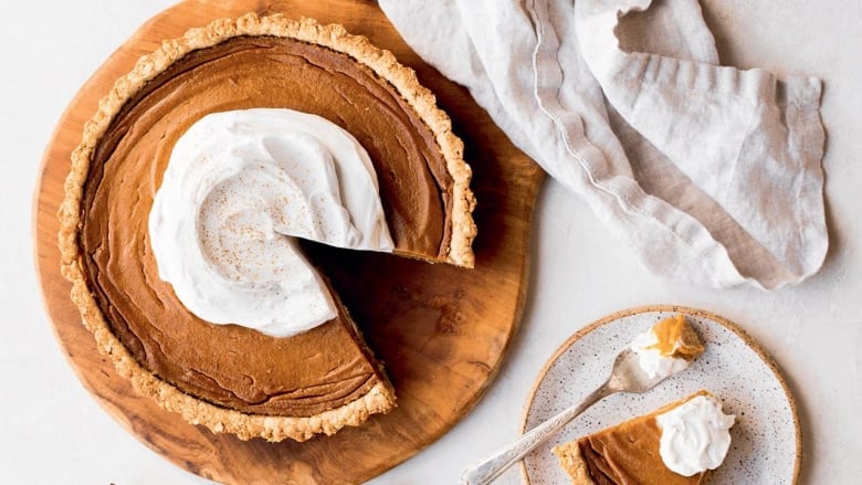 Overhead shot of a pie sitting on a wooden cutting board on a white surface. The pie is topped with whipped cream. A slice of it is on a white plate in the bottom right corner. 