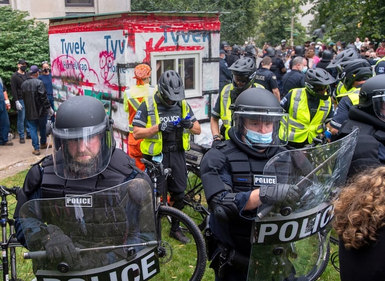 A line of police in riot gear surround a wooden shelter.