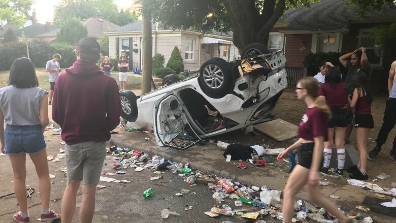 Young people stand around a car that has been flipped upside down on a sidewalk.