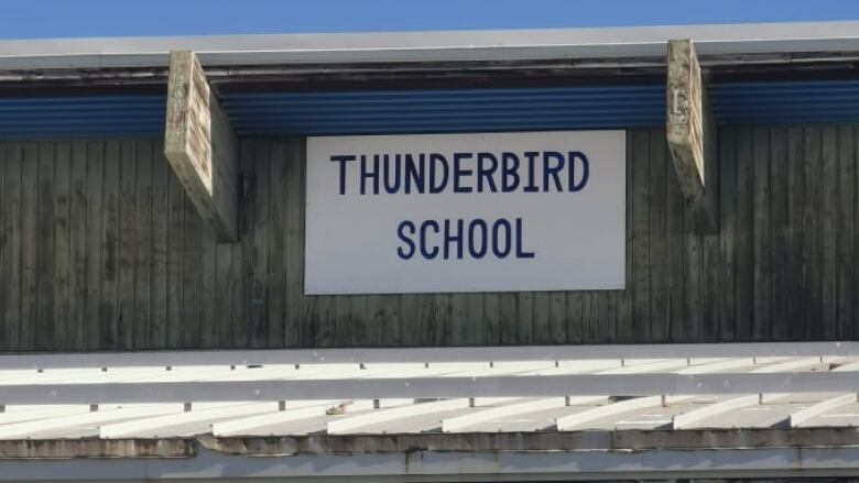 A white sign with blue letter says Thunderbrid School. It is attached to an outside wall, just below the overhang of the roof.