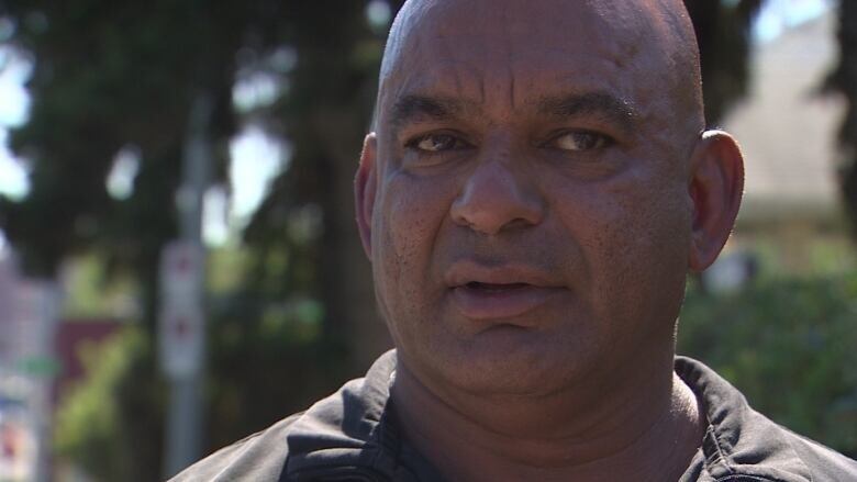 A man with a shaved head and a beige buttoned shirt looks away from the camera. 