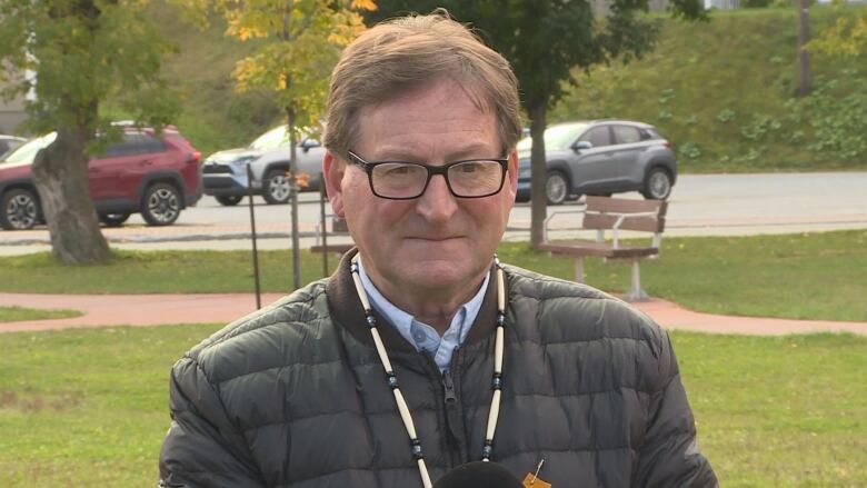 A man standing outside wearing a puffy jacket. He has brown-grey hair and black-rimmed glasses.