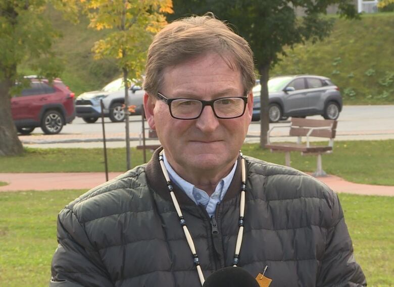 A man standing outside wearing a puffy jacket. He has brown-grey hair and black-rimmed glasses.