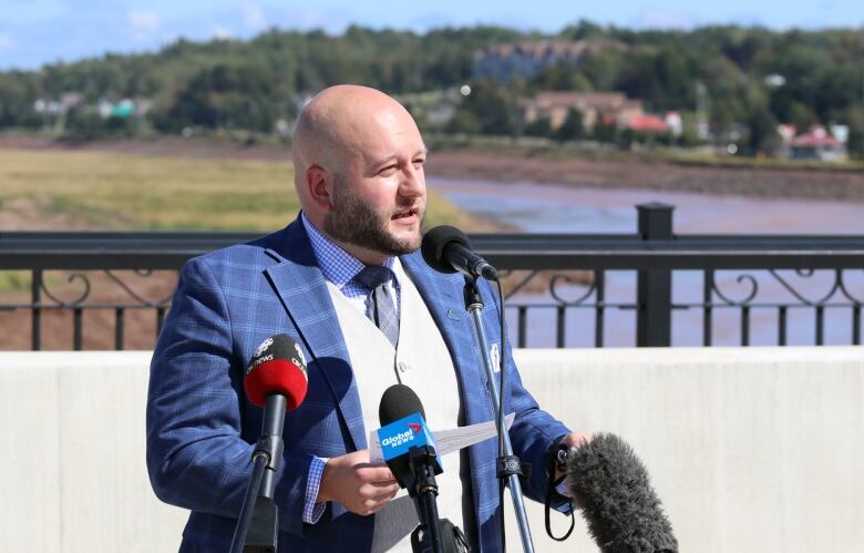 A bald man in blue suit stands outside on a bridge talking into microphones from news media.