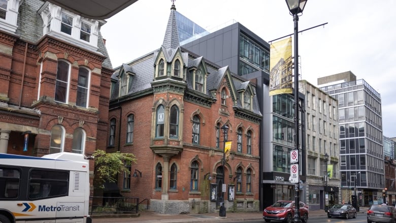 An older brick building with turret architecture is seen amongst glass commercial buildings on a street with cars.