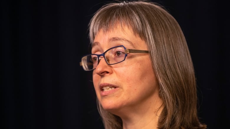 A woman in glasses speaking against a dark background.