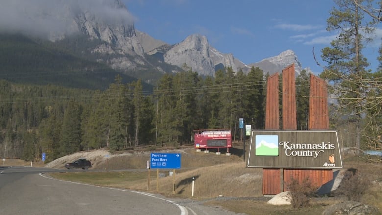 A sign that reads Kananaskis Country in front of a landscape of green trees, grey mountains and a blue sky.