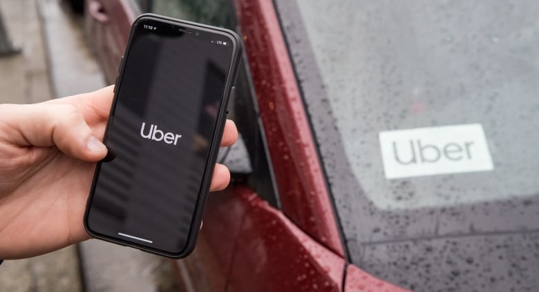 A hand holds an iPhone opened to the Uber app in front of the windshield of a car with an Uber sticker on it.