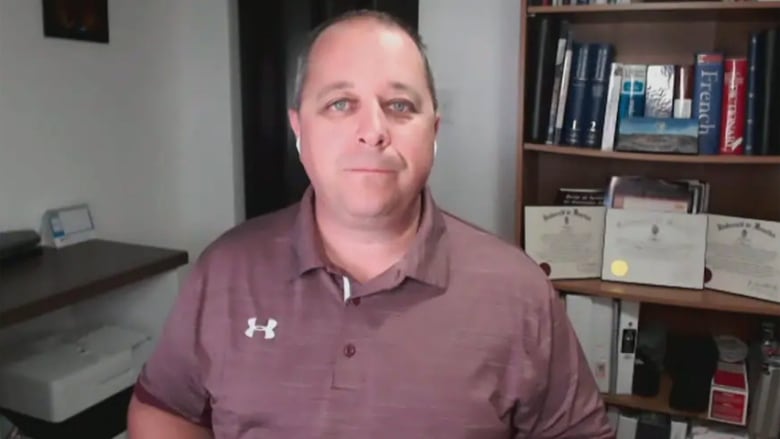 An unsmiling man wearing a muted red Under Armour polo shirt with a bookshelf behind him