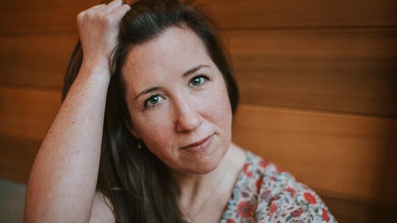 A portrait of a woman wearing a flowery shirt and with long dark hair that she is holding back from her face with her right hand