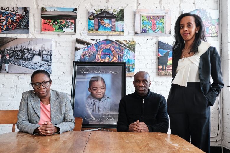 A man and a woman with serious expressions sit at a table, a framed picture of a child stands in the middle of them. Another woman stands on the side.