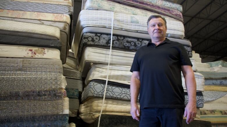 A man wearing a black polo shirt stands in front of a tall pile of mattresses inside a warehouse.