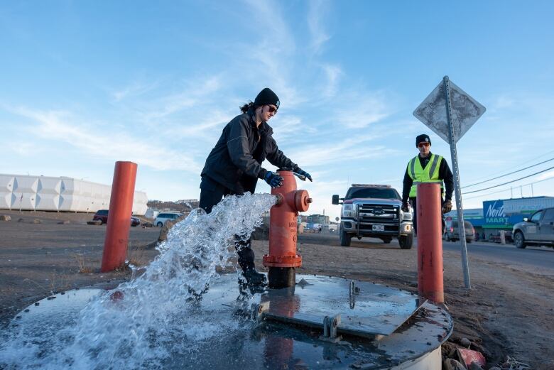 Two people stand by a fire hydrant as water pours out.