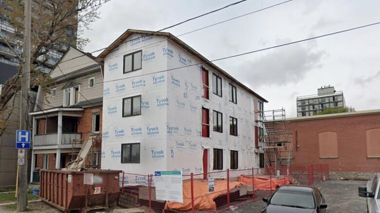 A low-rise four-unit apartment building is under construction on a city street, its sides still covered in white Tyvek sheeting.