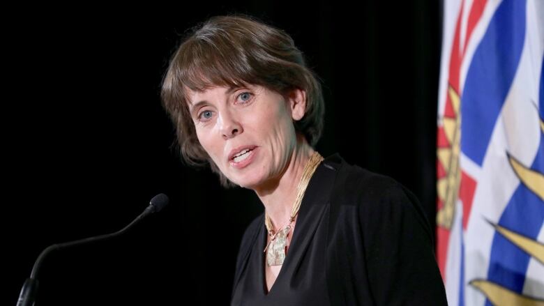 A white woman with chin-length brown hair is shown from the chest up, speaking into a microphone in front of a British Columbia flag.