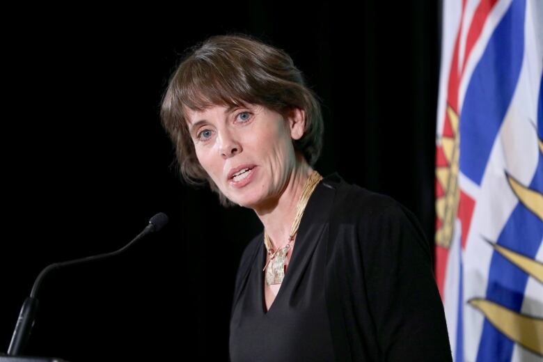 A white woman with chin-length brown hair is shown from the chest up, speaking into a microphone in front of a British Columbia flag.