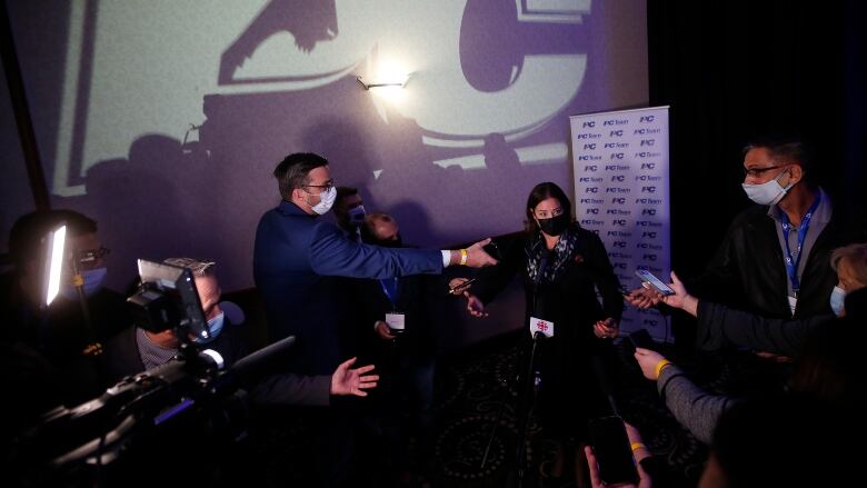 A PC logo is projected onto the wall above a scrum of reporters interviewing a woman.