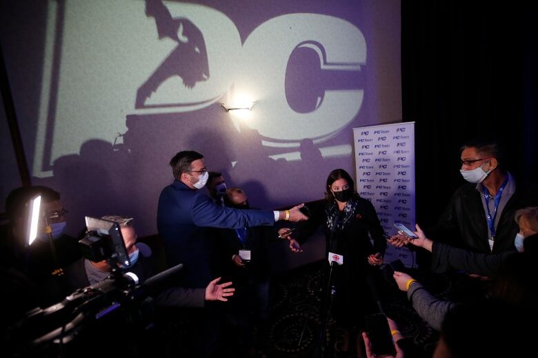 A PC logo is projected onto the wall above a scrum of reporters interviewing a woman.