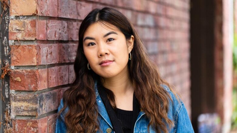 A young woman with long dark brown hair wearing a jean jacket over a black shirt and leaning against a brick wall