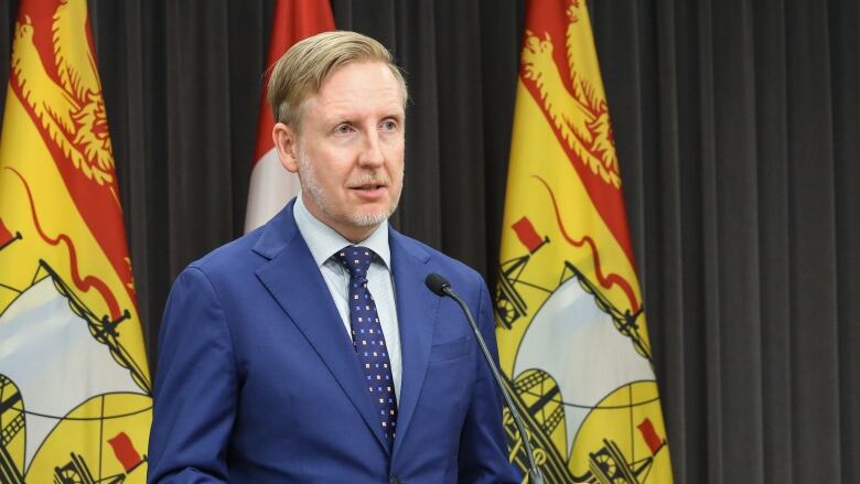 A man wearing a blue suit stands at a lectern. He is flanked by two New Brunswick flags. 