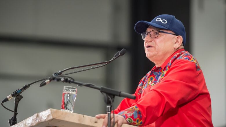 A man in a red shirt and a dark blue baseball cap stands at a podium.