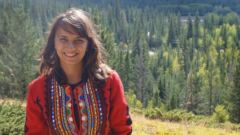 A smiling woman with dark brown hair wearing a colourful shirt with a forest in the background