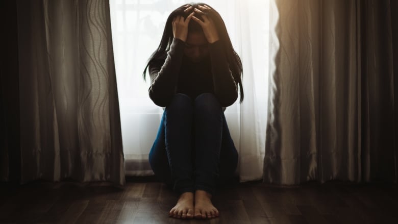 A silhouetted woman with long hair sits cross-legged with her head down and her hands on her head.