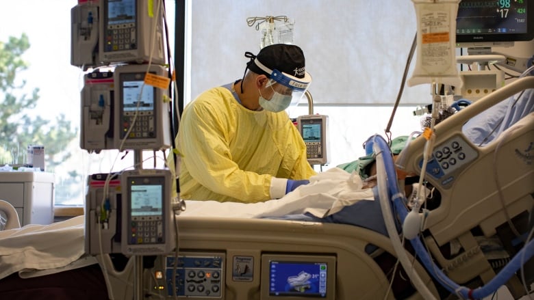 A nurse wearing PPE leans over a hospital bed with machines around it.