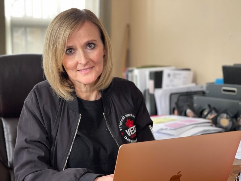 A woman with blonde hair and wearing a VETS Canada jacket sits at an Apple laptop.