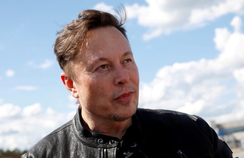 A man in a leather jacket stands outside with a blue sky and white clouds behind him.