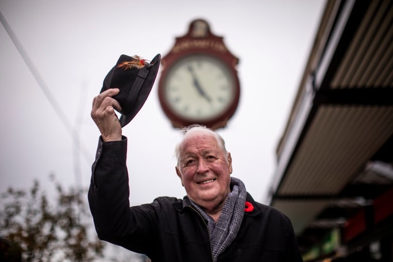 A man tips his hat in front of a giant clock taller than him in the blurred-out background.