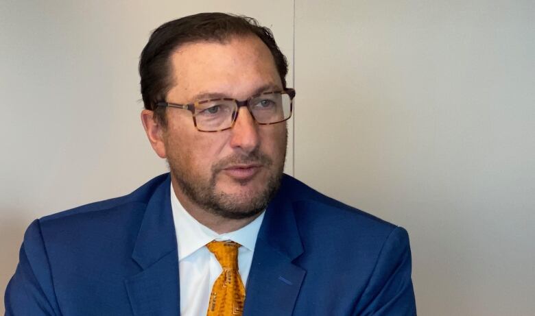 A man with short brown hair and glasses wears a blue suit with white shirt and orange tie. He is sitting.