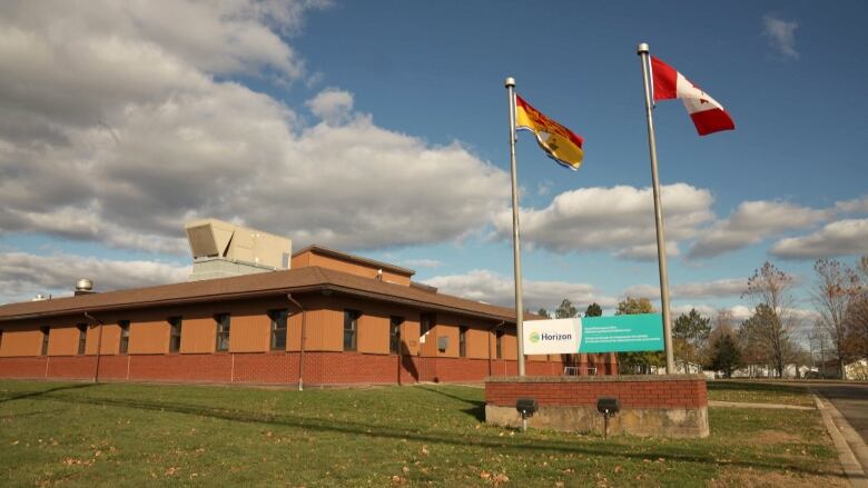 an exterior photo of a sand-coloured health facility with brick around the bottom half of the building. There is also a Horizon Health Network sign on the lawn in front of the building, with a New Brunswick flag and Canadian flag flying above and either side of the sign.