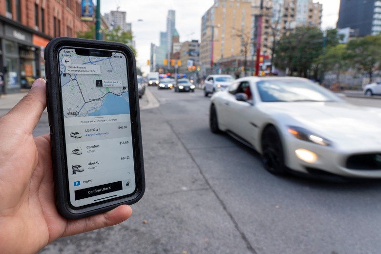 A car drives past a person holding out a phone displaying the Uber app on a busy street in downtown Toronto.  